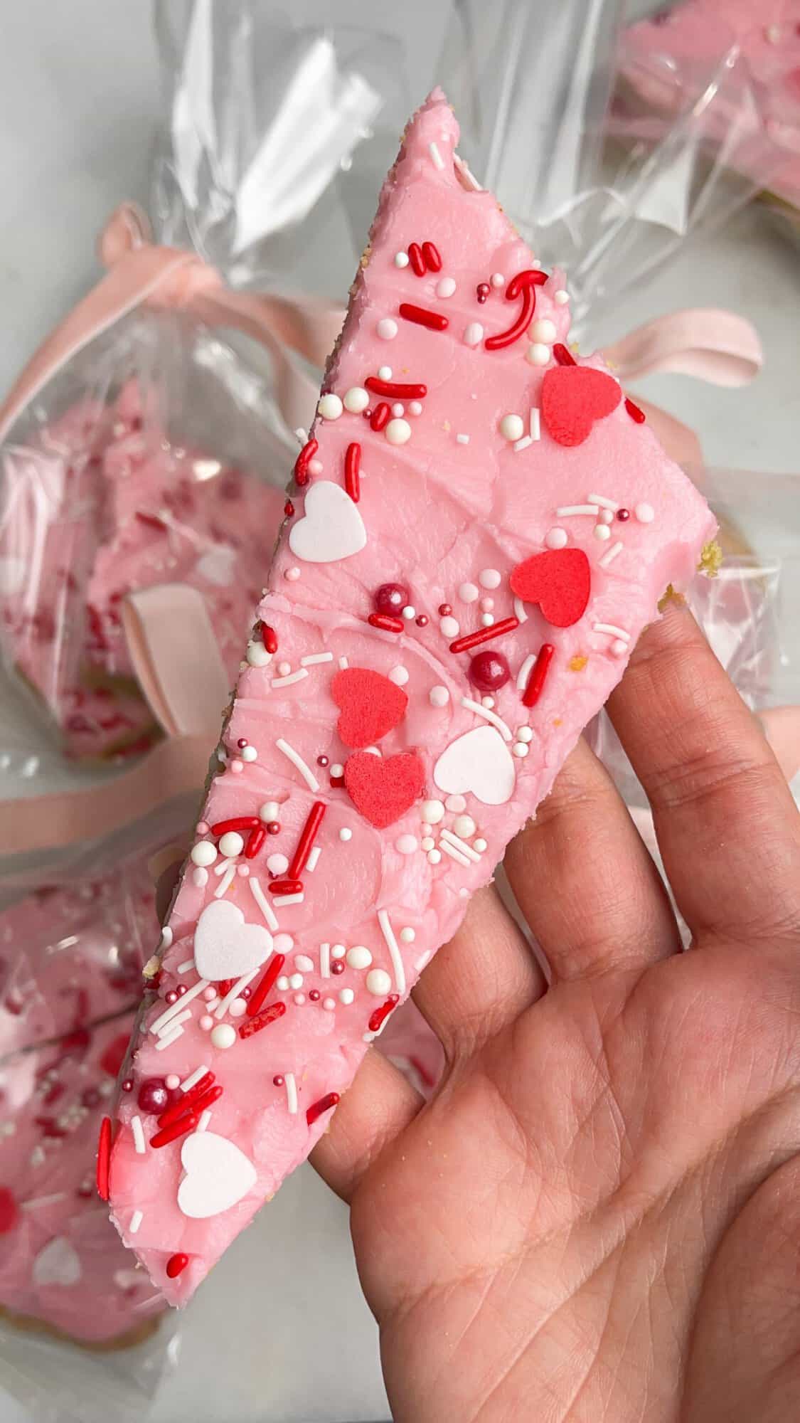 A thick, chewy cookie base is topped with a layer of sweet frosting and festive sprinkles in these frosted sugar cookie bars. A hand holding a frosted sugar cookie bar