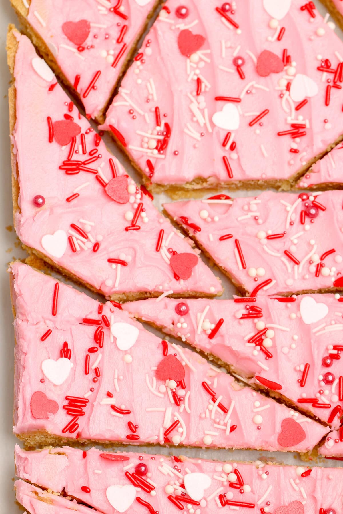 A thick, chewy cookie base is topped with a layer of sweet frosting and festive sprinkles in these frosted sugar cookie bars. Overhead view of frosted sugar cookie bars cut into various shapes
