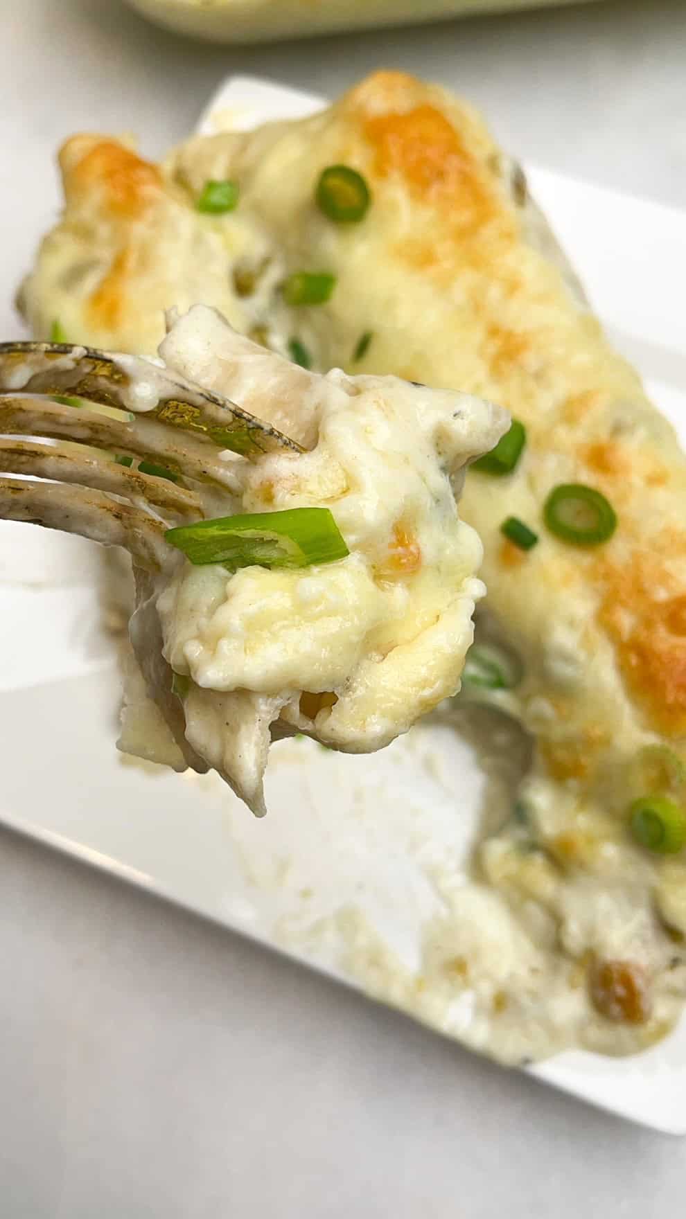 Seasoned chicken and shredded cheese are wrapped in tortillas and topped with a rich, tangy sauce in these White Chicken Enchiladas. A fork with a bite of white chicken enchilada