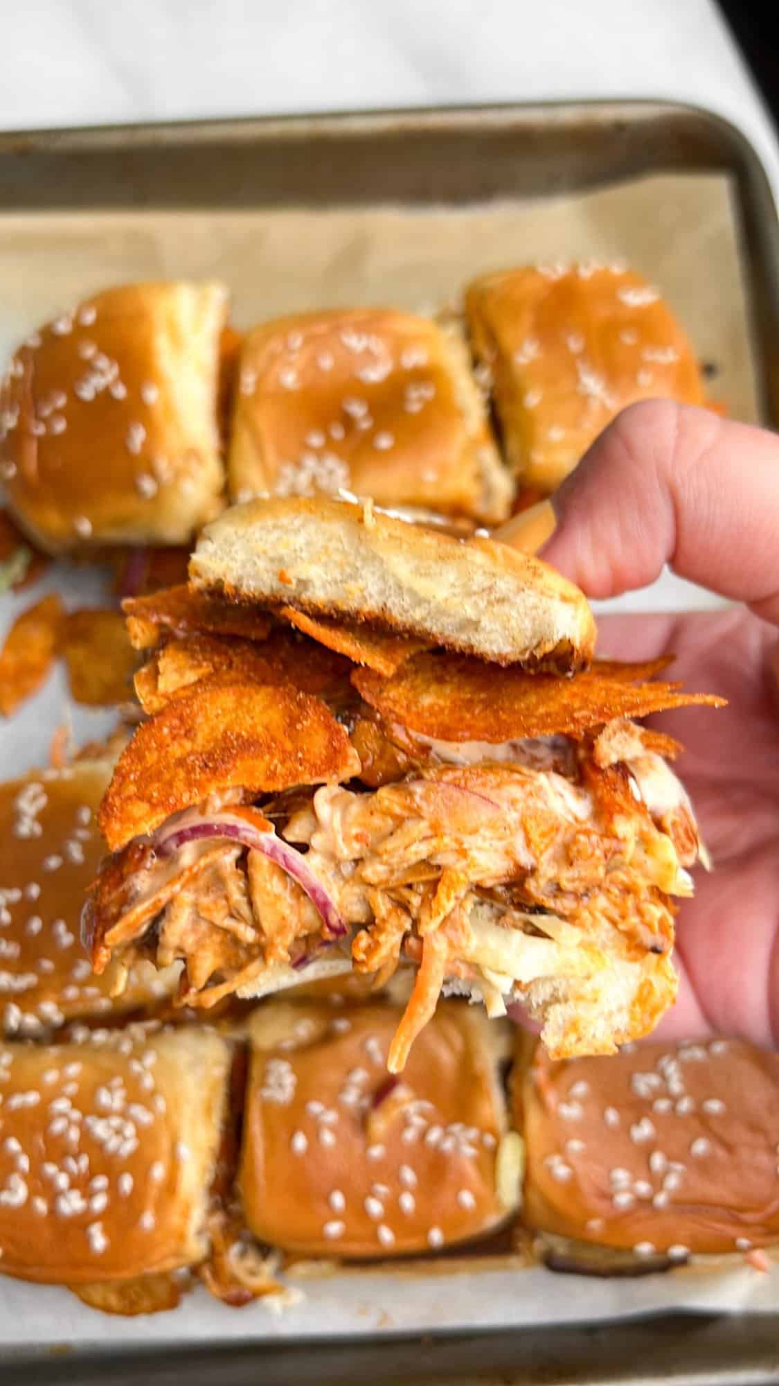 Shredded BBQ chicken, coleslaw tossed in Alabama white sauce, and crunchy chips make these BBQ Chicken Sliders unforgettable. A hand holding a BBQ chicken slider over the pan of sliders