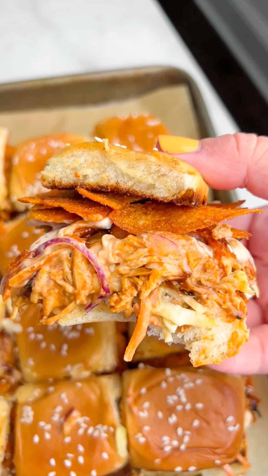 Shredded BBQ chicken, coleslaw tossed in Alabama white sauce, and crunchy chips make these BBQ Chicken Sliders unforgettable. A hand holding a BBQ Chicken Slider over a pan of sliders