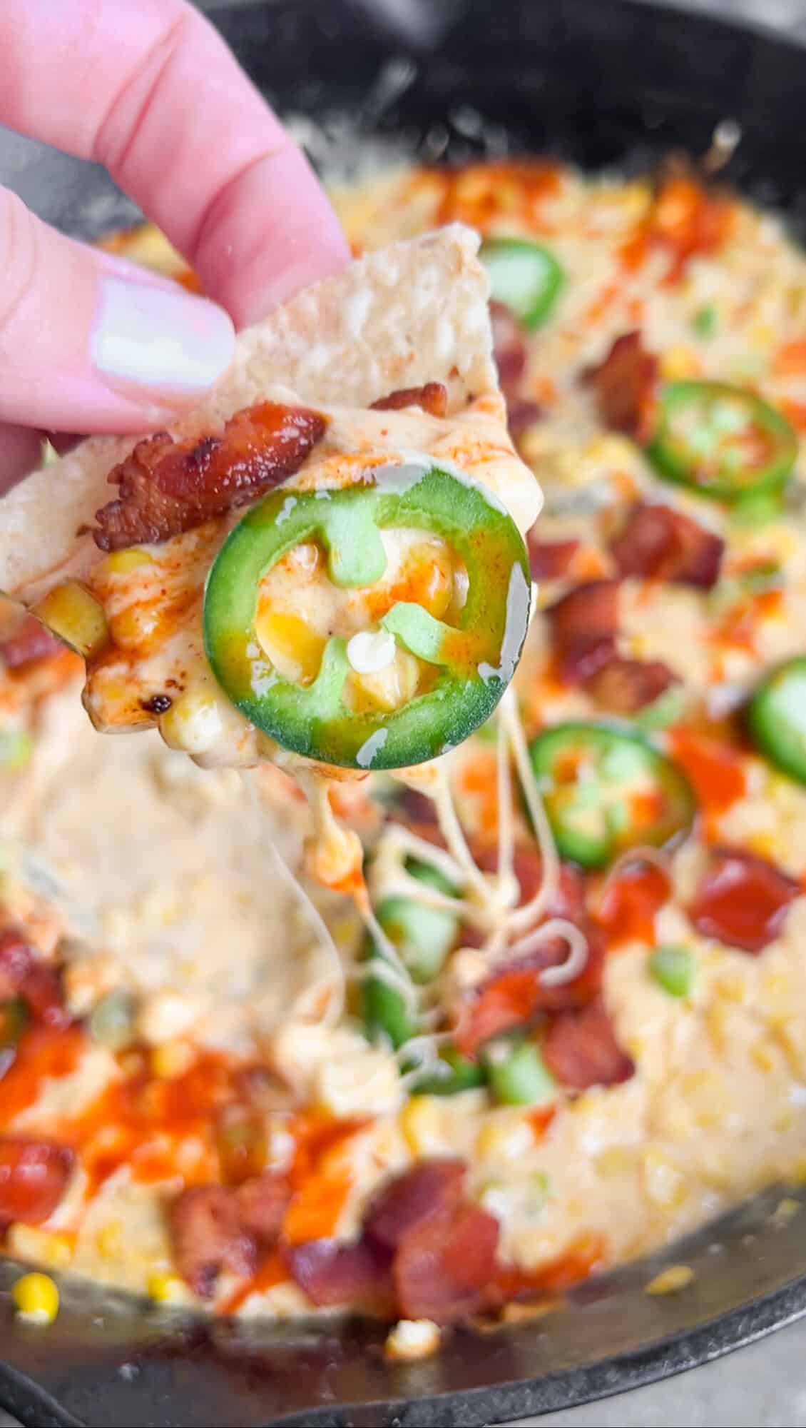This Hot Corn Dip is bubbling hot and loaded with kicks of heat and pops of sweet corn, all intertwined with gooey melty cheese. A hand holding a tortilla chip with hot corn dip