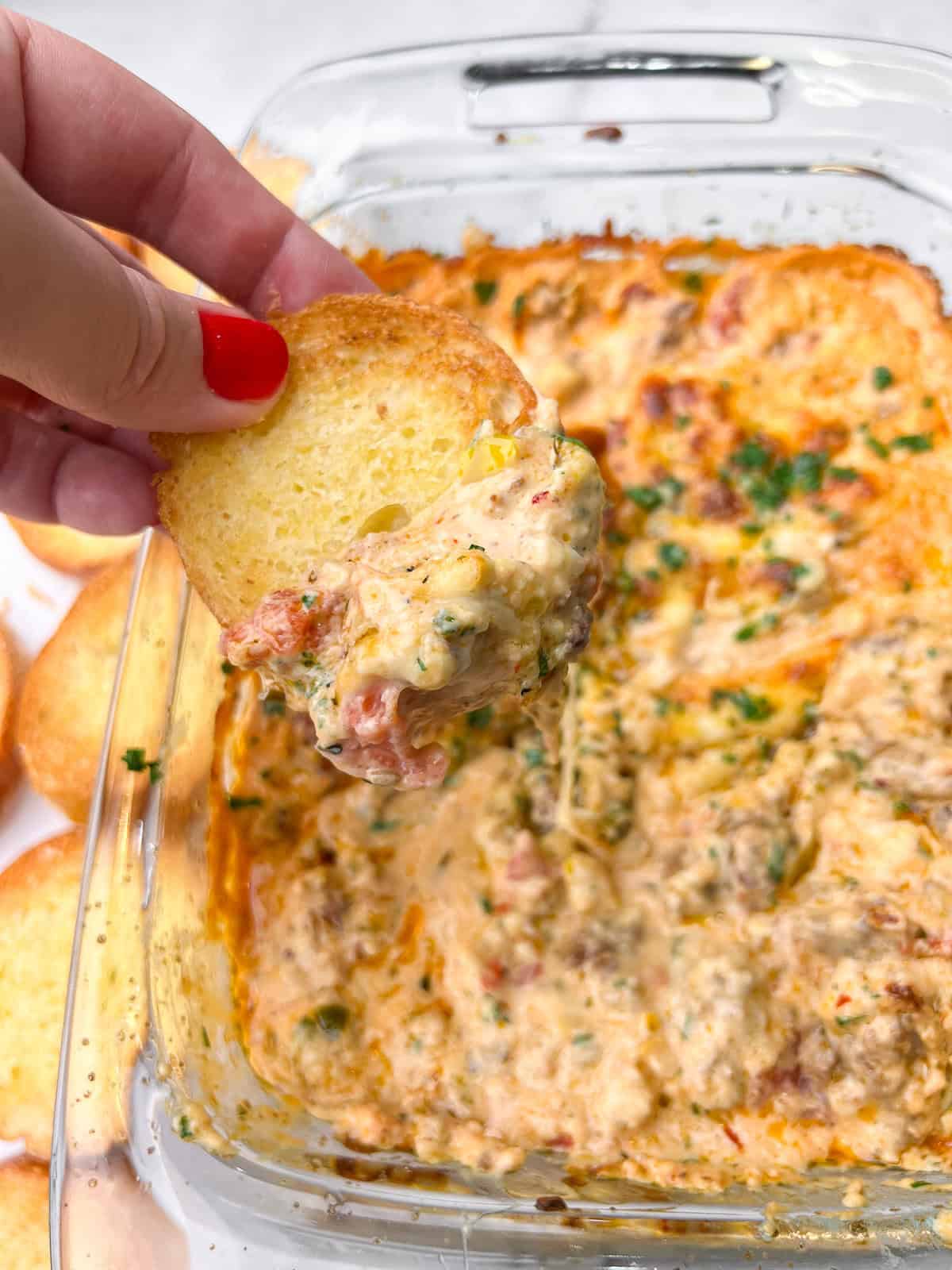 This 10-minute Spicy Italian Sausage Dip is made with mozzarella cheese, tangy pickled peppers, and hot sausage. Great for parties! A hand dipping a toasted crostini in spicy Italian sausage dip