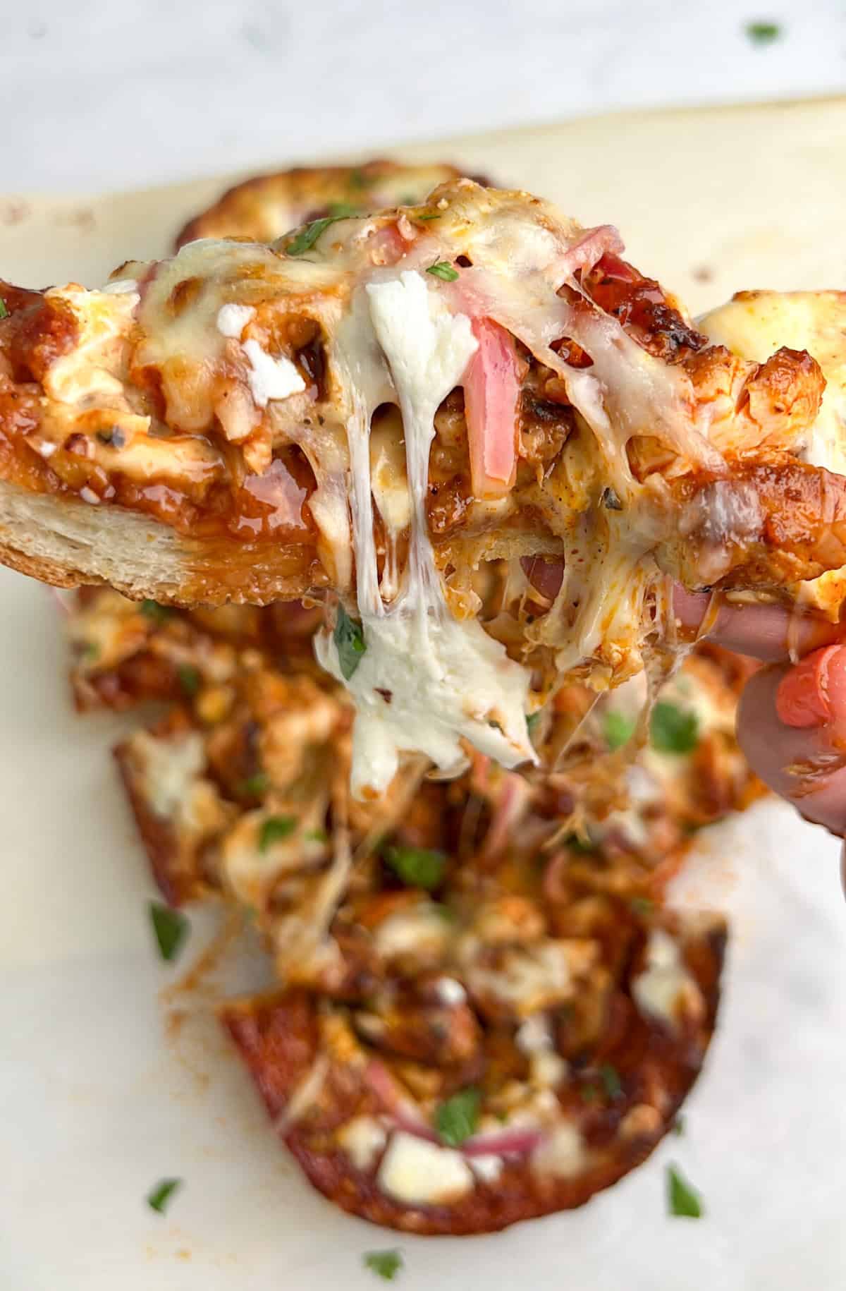 Crusty French bread is topped with sauce, cheese, and any toppings of choice, and then baked in this French Bread Pizza recipe. A hand holding a slice of BBQ chicken French bread pizza