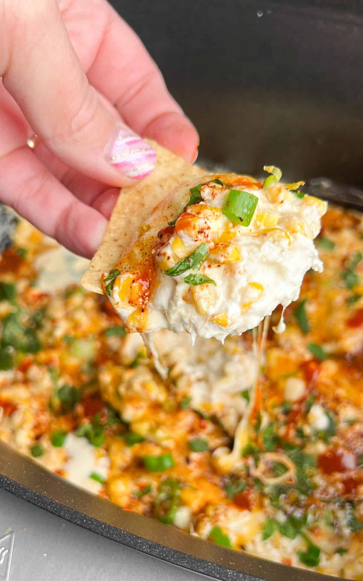 A hand holding a tortilla chip dipped in Mexican street corn dip