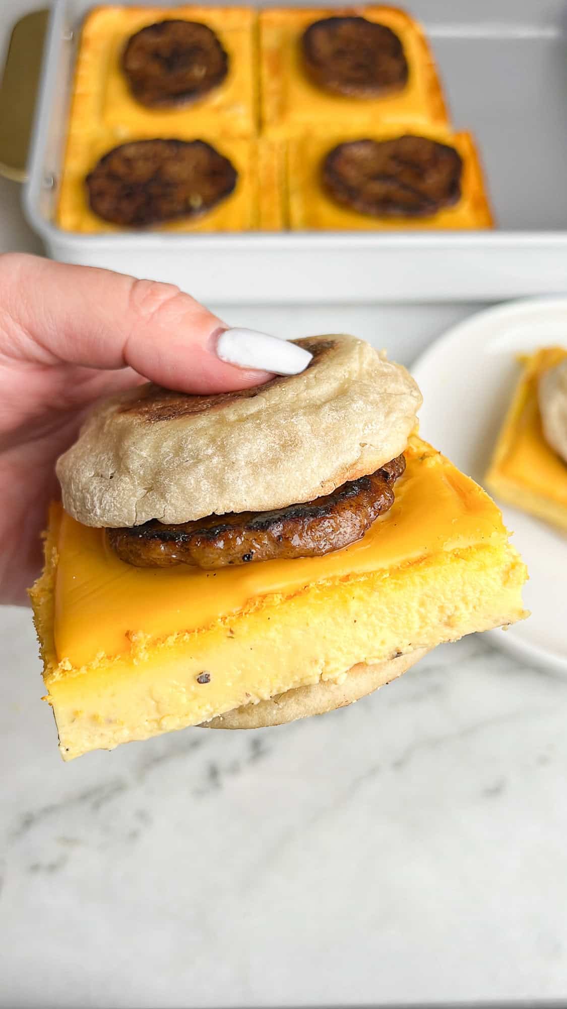 These Sheet Pan Breakfast Sandwiches are made with soft, custard-like eggs, sharp cheddar & sausage patties, on a buttery English muffin. A hand holding a sausage egg and cheese sheet pan breakfast sandwich