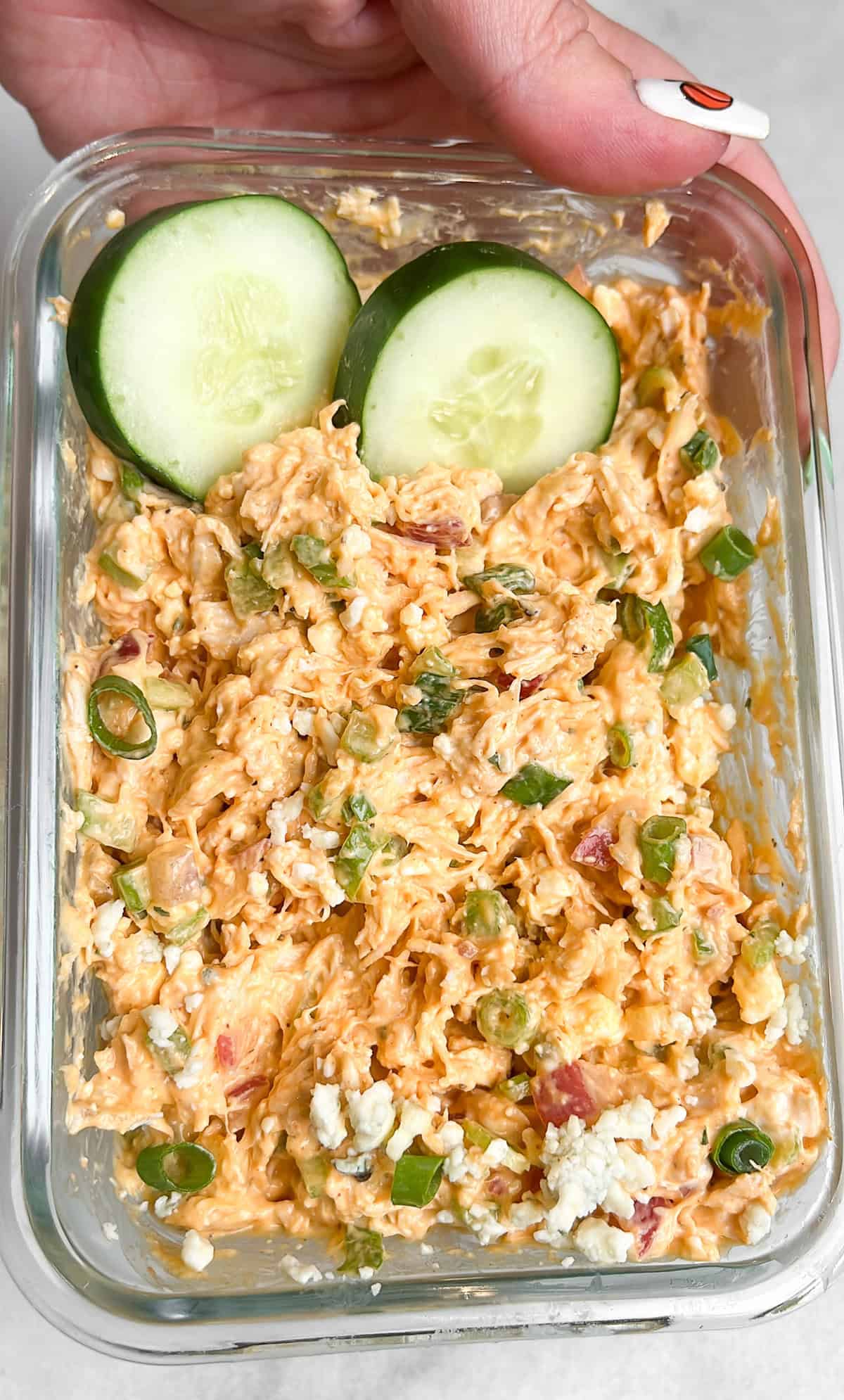 Made with rotisserie chicken and a Greek yogurt dressing, this Buffalo Chicken Salad is an easy, filling 10-minute recipe. A hand holding a container of buffalo chicken salad with slices of cucumber