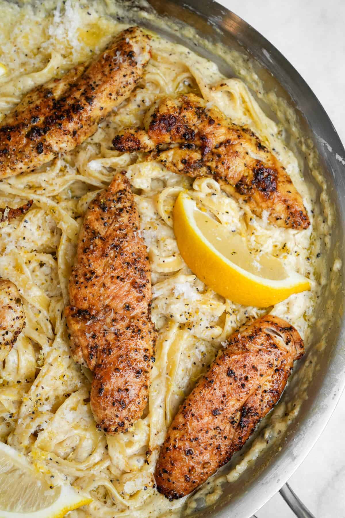 lemon pepper chicken on fettuccine alfredo in a pan with lemon wedges