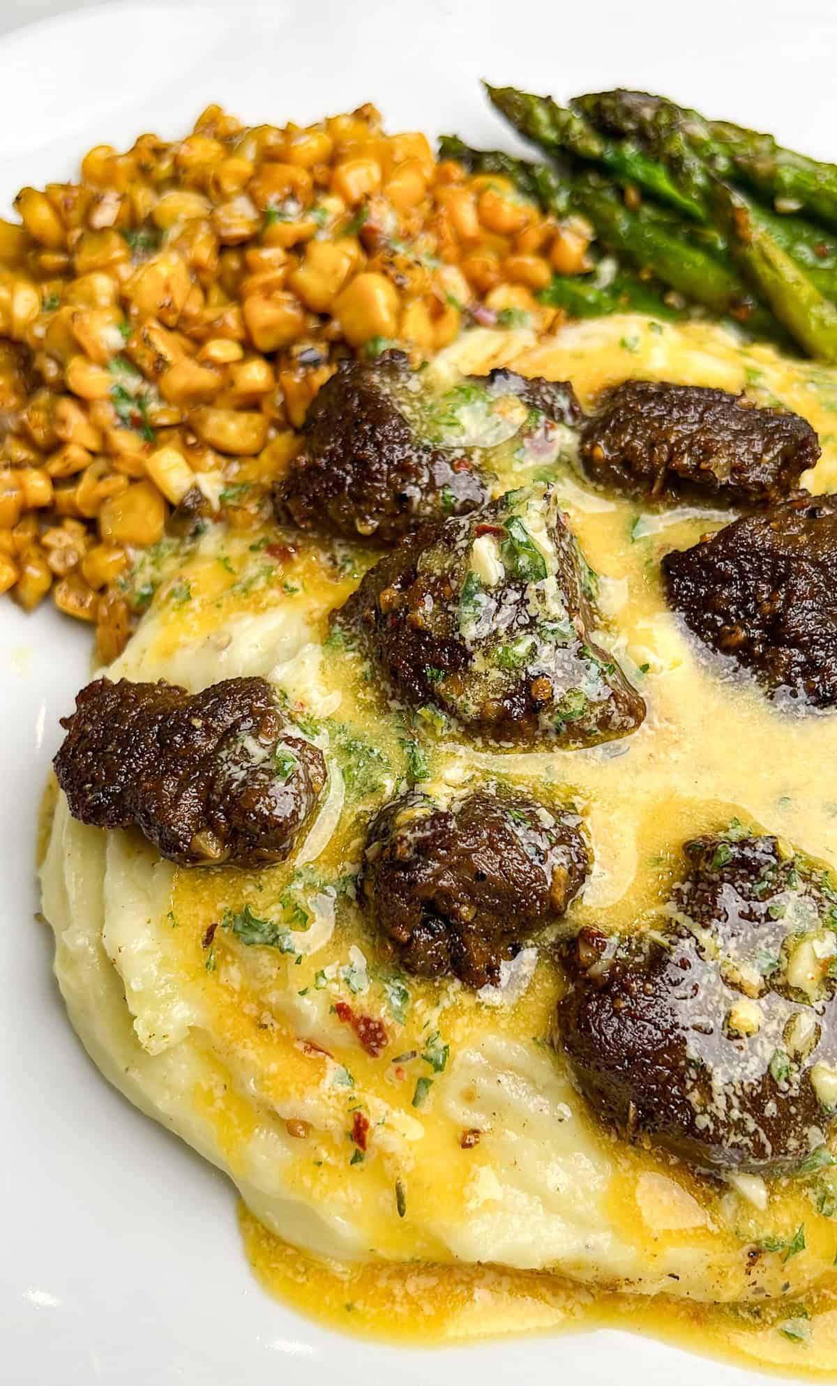 With steak bites served over mashed potatoes and drizzled with cowboy butter, this steak and potatoes recipe is perfect for any occasion. A dinner plate with steak and potatoes, asparagus and corn drizzled with cowboy butter