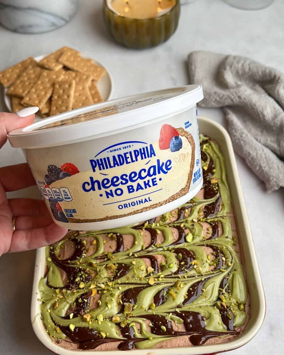 With layers of crispy kataifi, pistachio cream, and chocolate whipped cream, this Dubai chocolate cheesecake is a dream come true. A hand holding no-bake Philadelphia cheesecake spread up to the camera.