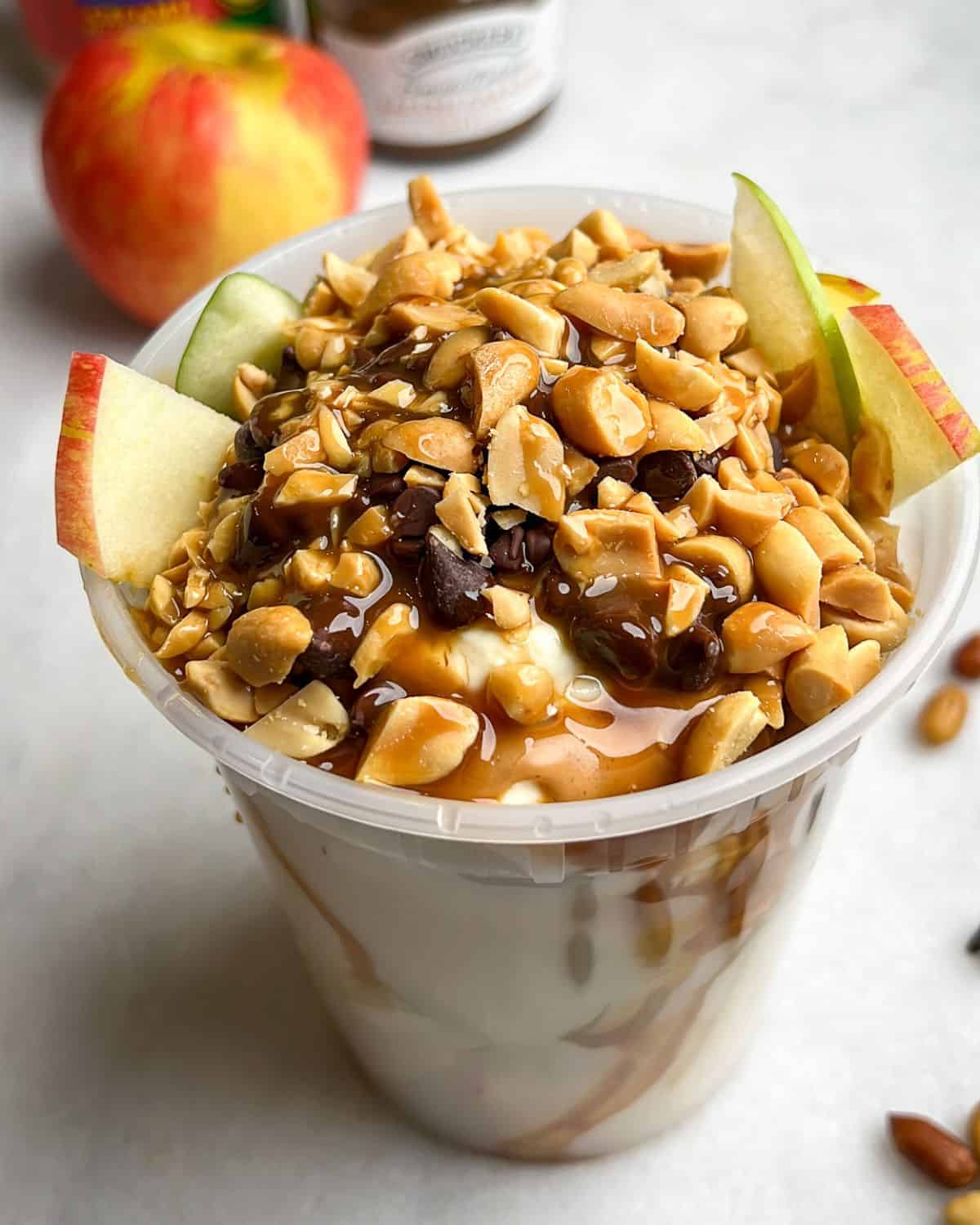 apples in a container with  peanut butter, caramel, and greek yogurt. Topped with peanuts and chocolate chips