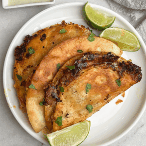 Three crispy beef tacos on a white plate with lime wedges