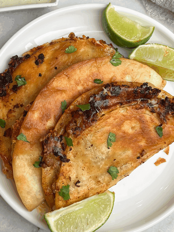 Three crispy beef tacos on a white plate with lime wedges