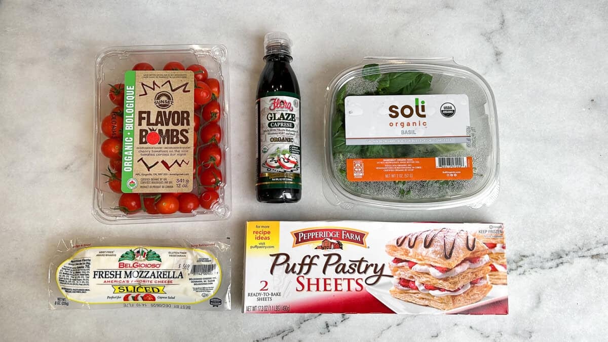 These easy caprese bites combine the fresh flavors of cherry tomatoes, mozzarella, and basil in a warm, flaky crust. Overhead view of cherry tomatoes, balsamic glaze, basil, mozzarella cheese, and puff pastry on the counter