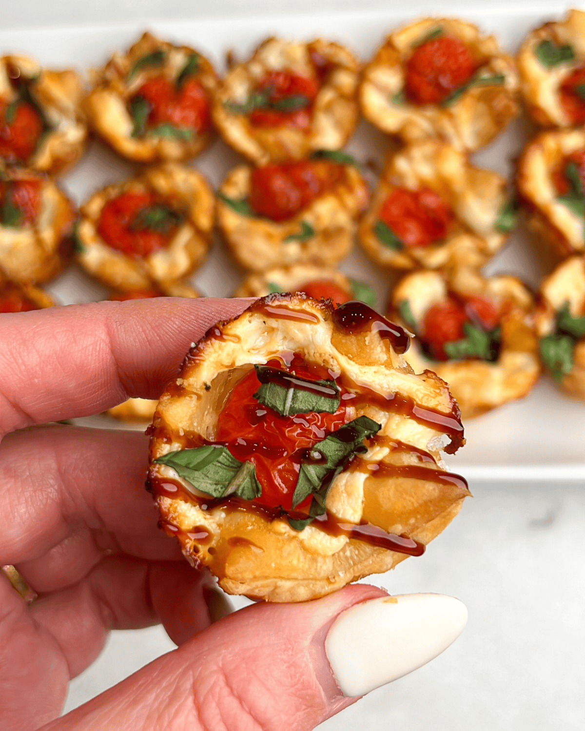 These easy caprese bites combine the fresh flavors of cherry tomatoes, mozzarella, and basil in a warm, flaky crust. A hand holding a caprese bite