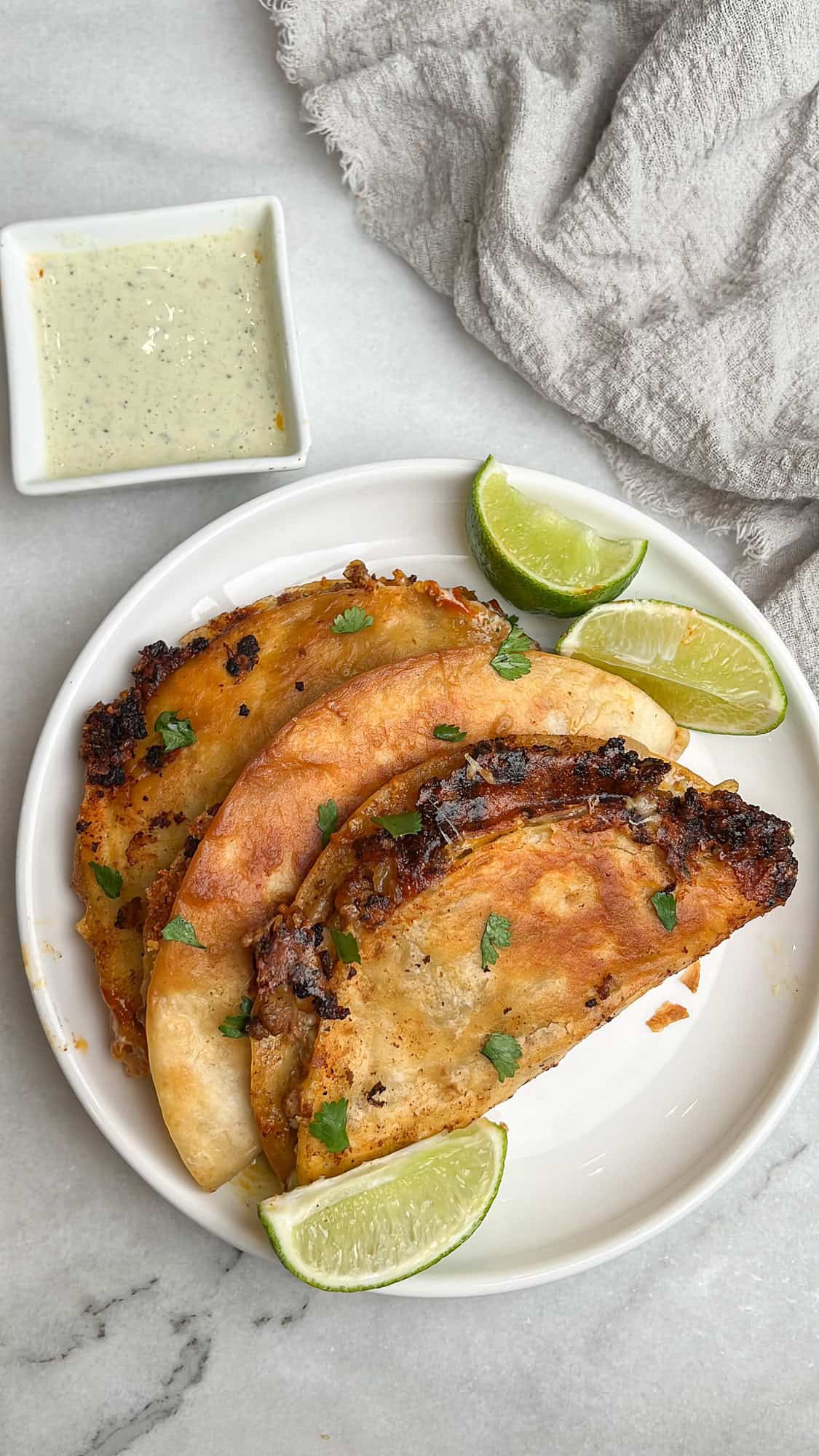 These Crispy Beef Tacos are loaded with savory, seasoned ground beef and lots of cheese, then quickly fried to crispy, melty perfection. Overhead view of three crispy beef tacos on a white plate with lime wedges