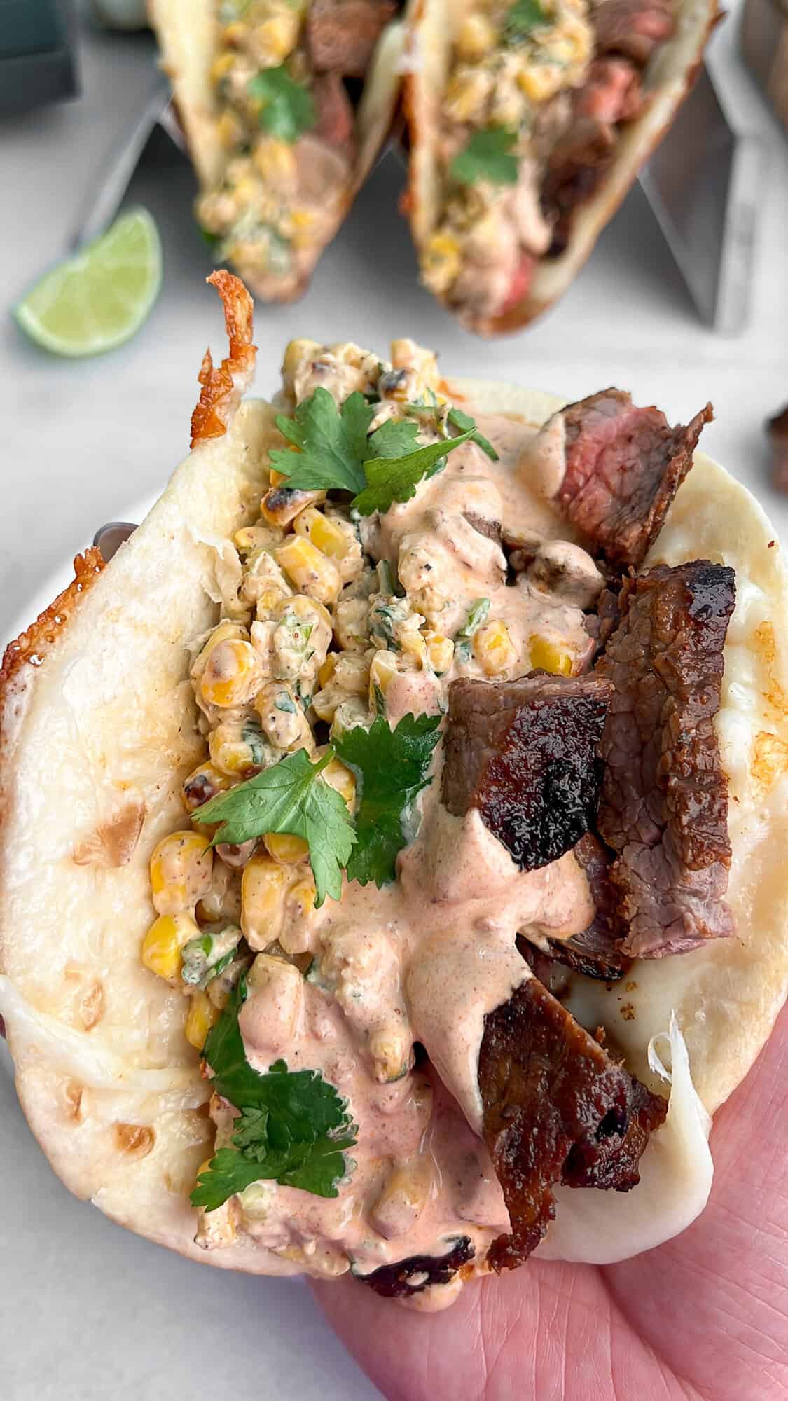 This Carne Asada Tacos recipe features juicy, citrus-marinated flank steak topped with street corn salad in cheesy tortillas. A carne asada taco topped with street corn salad and chipotle crema