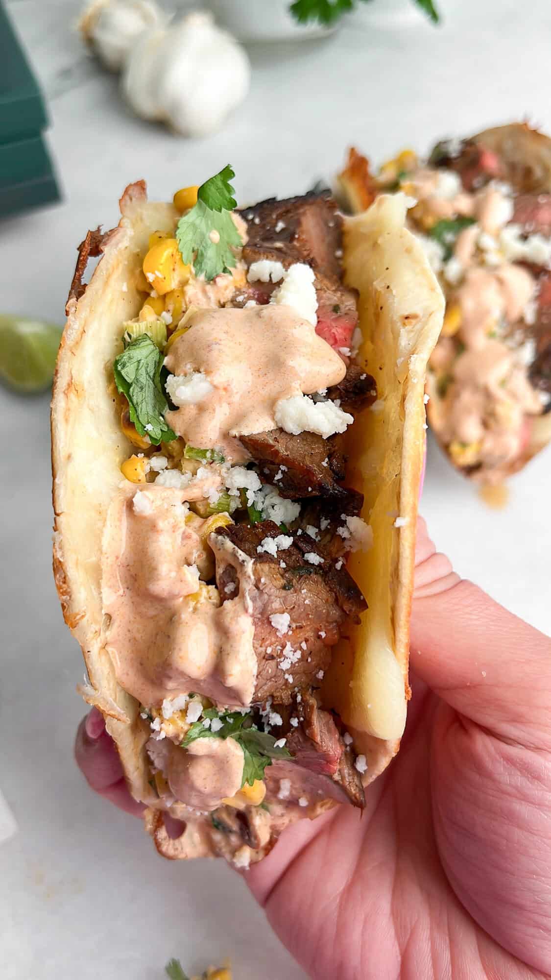 This Carne Asada Tacos recipe features juicy, citrus-marinated flank steak topped with street corn salad in cheesy tortillas. A hand holding a carne asada taco