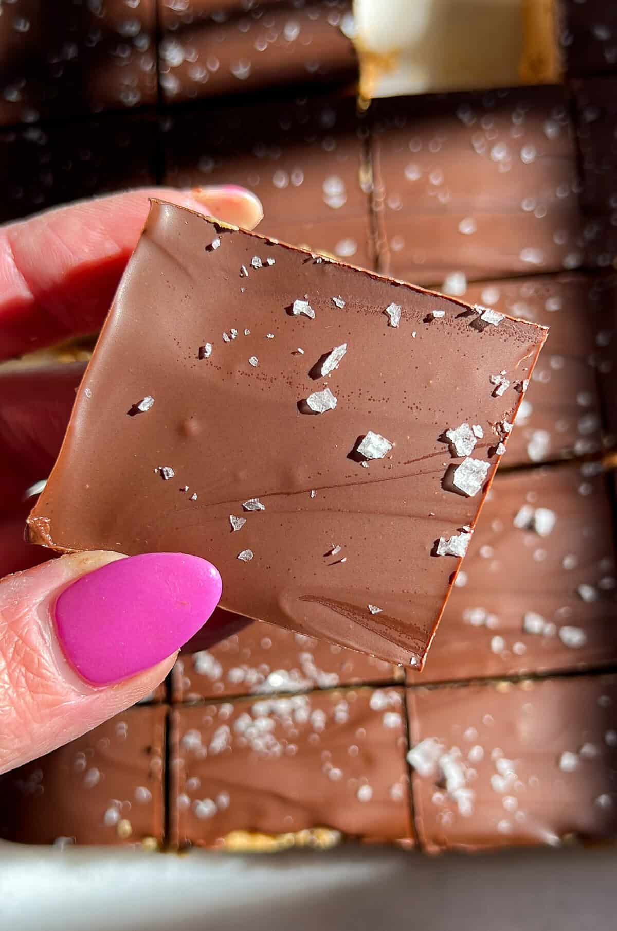 These no-bake peanut butter bars have a soft, sweet peanut butter base and a rich chocolate top with a sprinkle of flaky salt. A hand holding a chocolate peanut butter bar