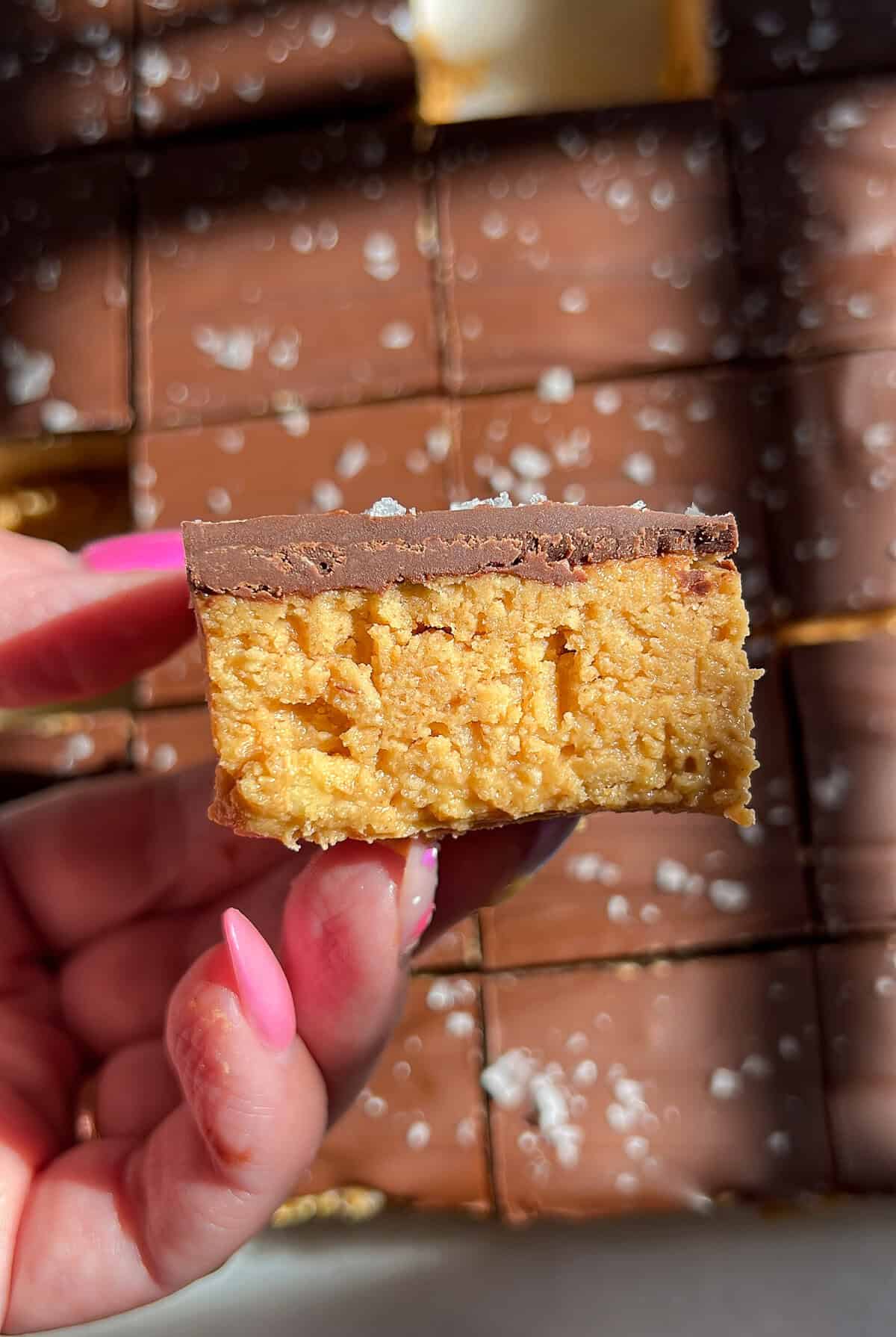 These no-bake peanut butter bars have a soft, sweet peanut butter base and a rich chocolate top with a sprinkle of flaky salt. A hand holding a no-bake peanut butter bar on its side showing the layers