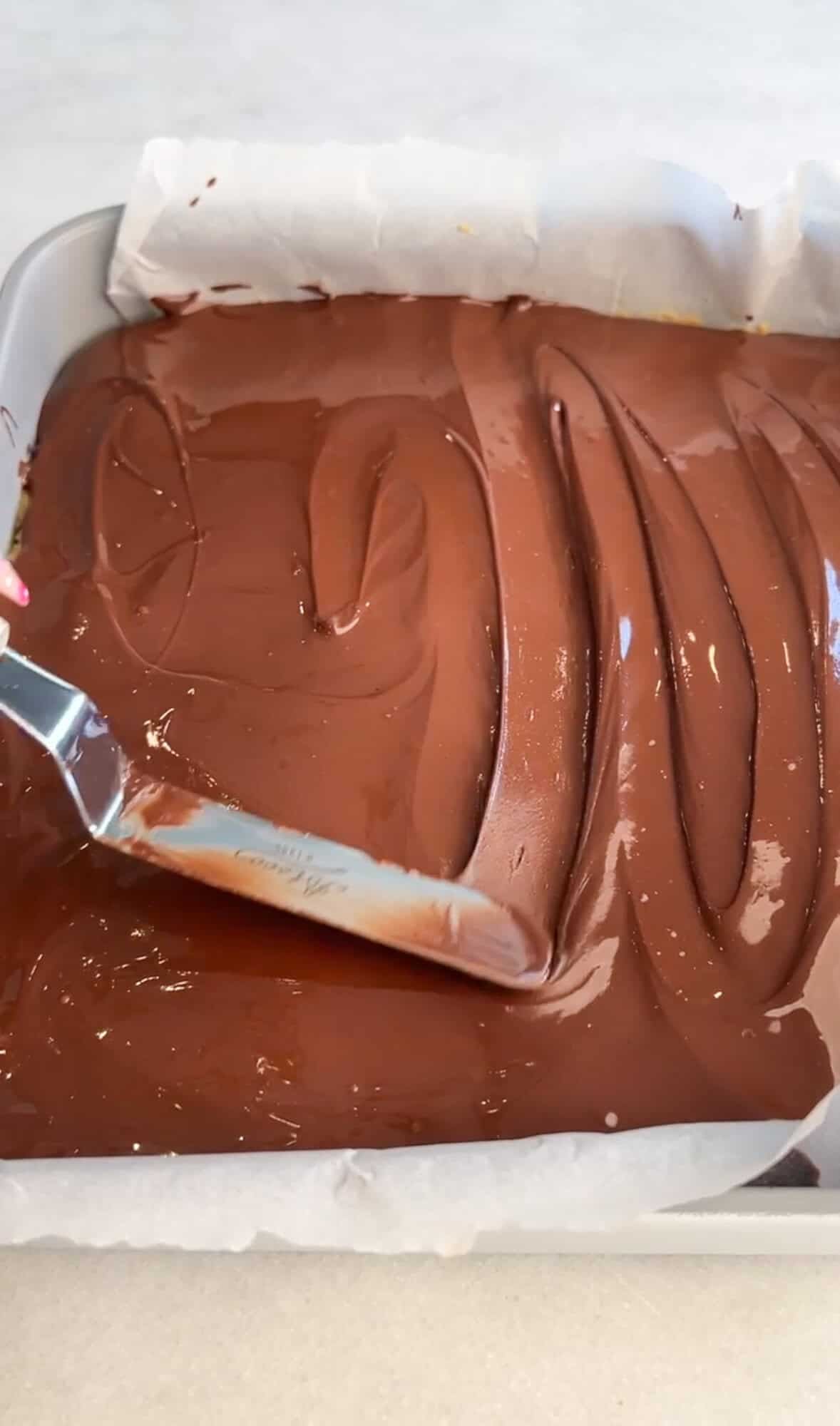 These no-bake peanut butter bars have a soft, sweet peanut butter base and a rich chocolate top with a sprinkle of flaky salt. Spreading the chocolate layer with a knife.