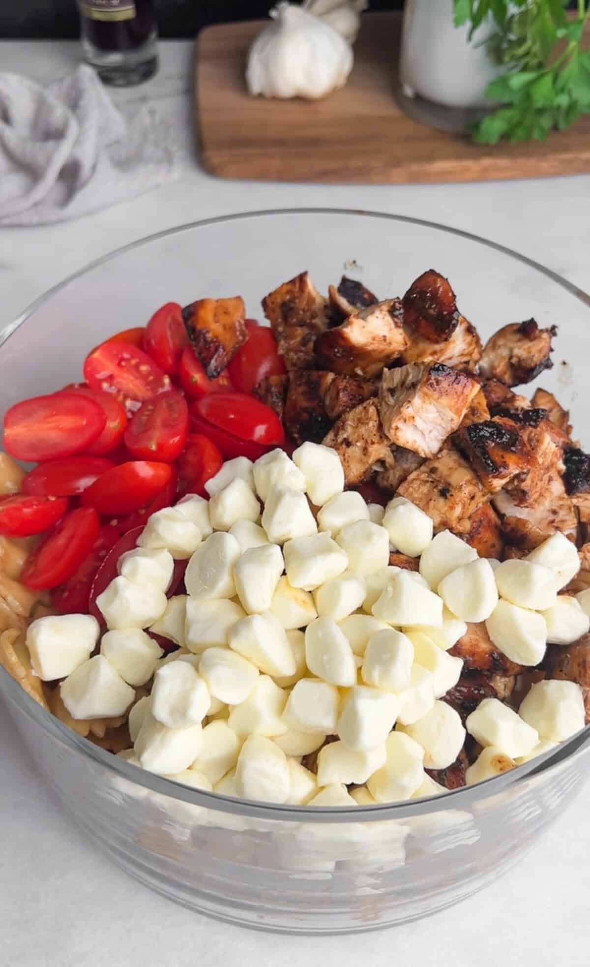 Caprese Pasta Salad is loaded with marinated chicken, tomatoes, mozzarella pearls, bacon, and basil tossed in a tangy balsamic dressing. Grape tomatoes, chicken, and mozzarella pearls in a glass bowl